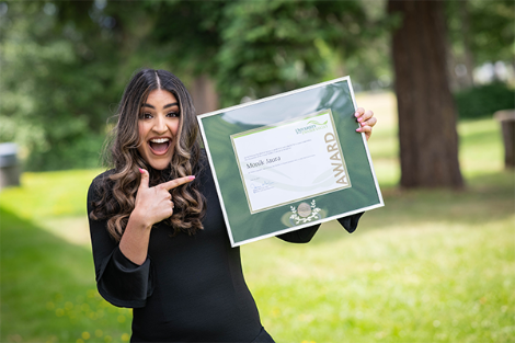 Smiling young woman pointing to a framed award she is holding up in one hand.