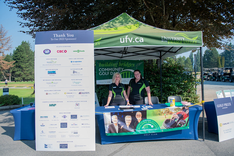 Two individuals staffing an informational booth with a banner listing sponsors, under a branded tent.
