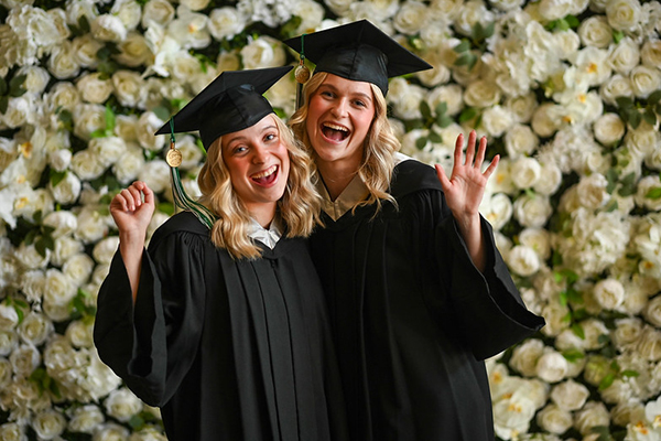 Two people in graduation gowns and caps cheerfully pose in front of a flower wall, waving and smiling at the camera.