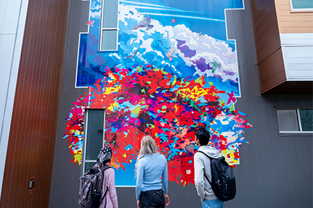 Three people with backpacks stand in front of a colorful, abstract mural on a building wall.