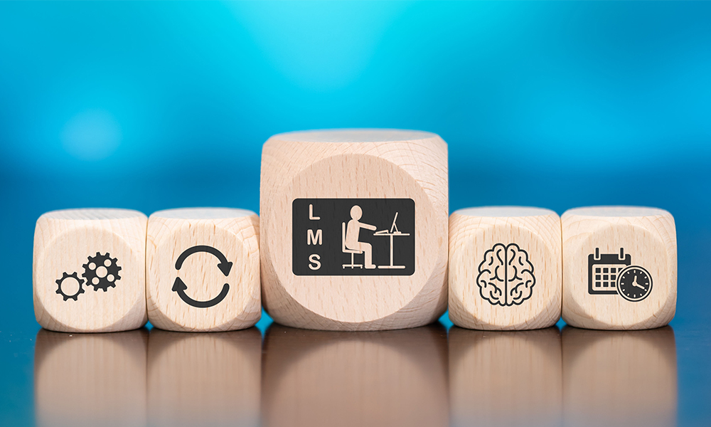 Five wooden blocks with icons: gears, refresh symbol, person at desk labeled LMS, brain, and calendar with clock, set against a blue background.