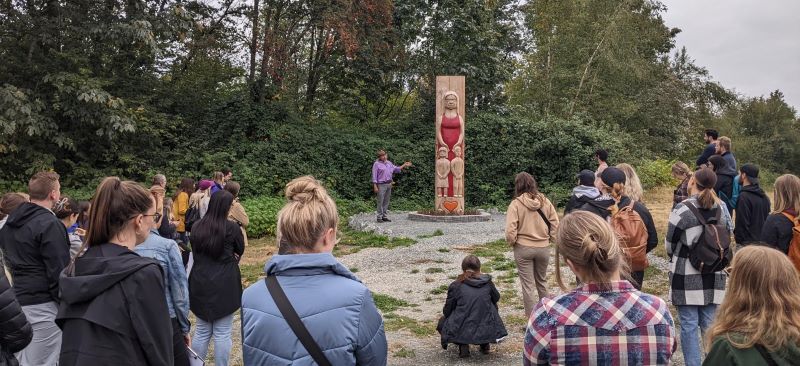 People gather around an Indigenous teacher who's pointing to a totem pole